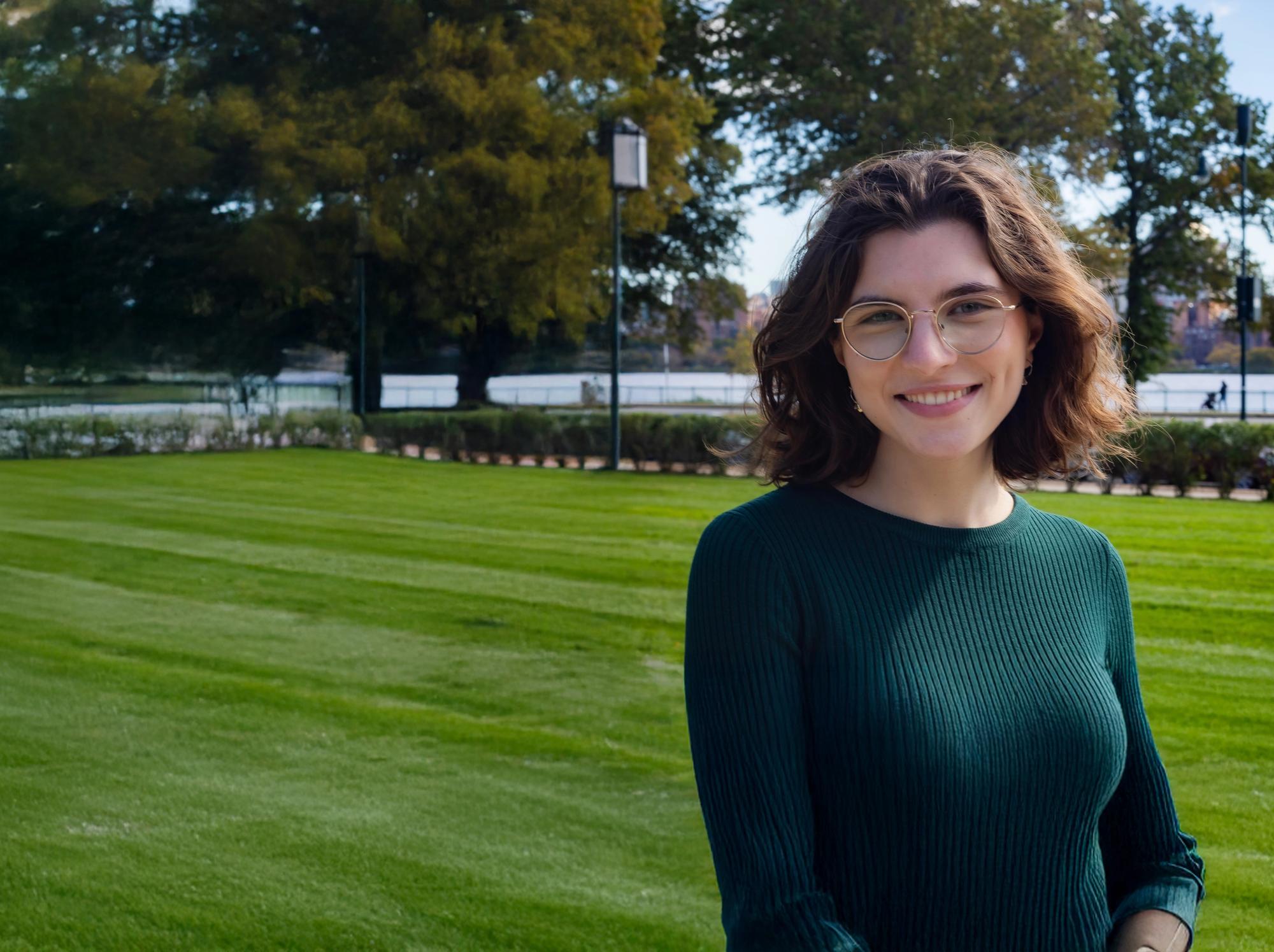 Young woman with shoulder-length curly hair and glasses smiling outdoors on a sunny day. She is wearing a dark green ribbed sweater and standing on a well-manicured lawn, with trees, light posts, and a river visible in the background.